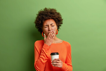 Photo of beautiful tired woman yawns and being sleepy after awakening early in morning holds disposable cup of coffee dressed in casual jumper isolated on vivid green background. Exhausted girl - obrazy, fototapety, plakaty