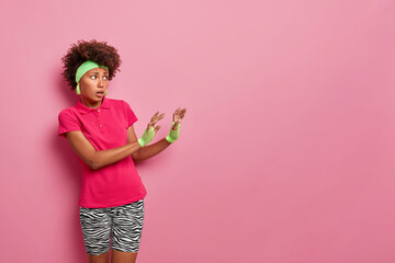 Displeased dark skinned young Afro American woman keeps hands towards empty space and asks not come closer sees something unpleasant wears headband t shirt leggings poses against pink background