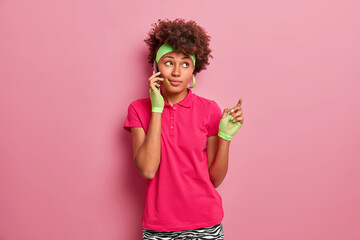 Pretty thoughtful Afro American woman with dark skin speaks on mobile phone spends free time indoors calls to friend dressed in active wear poses against pink background looks away pensively