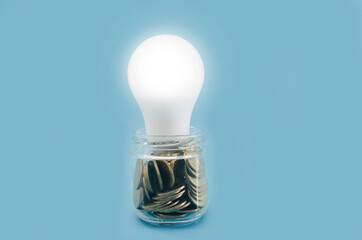a jar of coins and a lit energy-saving light bulb. Blue background. Electricity saving concept.
