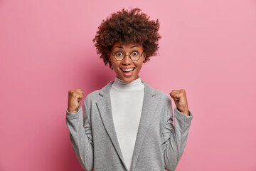 Fototapeta na wymiar Optimistic excited businesswoman raises clenched fists and celebrates triumph or successful project fulfillment dressed in formal grey clothes wears round spectacles isolated on pink studio wall