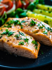Fried salmon fillet with boiled potatoes, lemon and vegetable salad served on black plate on wooden table

