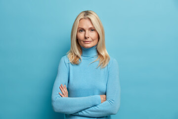 Self confident serious calm middle aged experienced blonde woman stands with arms folded dressed in casual turtleneck shows professional vibe stands in assertive pose against blue background.