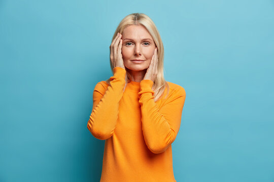 Beauty aging and cosmetology concept. Good looking serious woman with blonde hair touches face gently has healthy skin after applying anti aging cream wears bright sweater isolated on blue wall