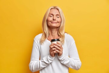 Calm relaxed senior woman holds hot beverage in disposable cup stands closed eyes in casual wear isolated on yellow background. Tranquil satisfied middle aged lady enjoys free time as drinks espresso