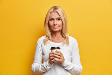 Fototapeta premium Studio shot of serious blonde woman holds disposable cup of coffee looks confident at camera has wrinkled skin wears casual white jumper enjoys free time. Middle aged lady poses with hot drink