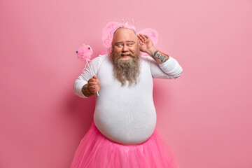 Funny overweight bearded man dressed in fairy costume to entertain guests of party keeps hand near ear and tries to overhear something holds magic wand has big belly poses against pink background