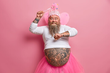 Happy amused bearded European man has fun on carnival or costume party dances with raised arms holds magic wand wears fairy outfit has fat tattooed belly. People holiday celebration concept. - obrazy, fototapety, plakaty