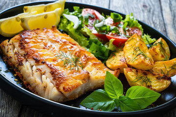 Fish dish - fried cod fillet with potatoes and vegetable salad on wooden table
