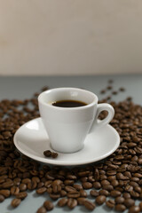White cup with espresso on a white saucer among coffee beans on a gray background. Copy space. Selective focus. Vertical orientation.