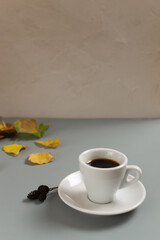 White cup with espresso on a white saucer among autumn yellow and green leaves on a gray background. Copy space. Selective focus. Vertical orientation.