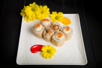 Side view of Asian traditional Ebi Tempura Sushi Roll pieces with tempura shrimp wrapped in rice and sesame seeds served on white Japanese plate with yellow camomile flowers and paining brush strokes
