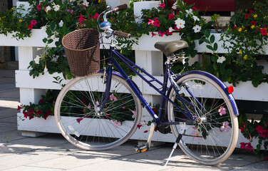 Bicicleta azul com cesto na frente parada junto de uma vedação de madeira com flores roxas e brancas