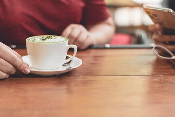 Hot green tea cup​ on wood table and two women meeting use smartphone in a morning at the cafe shop, Cozy atmosphere is for relaxing on vacation or charge energy to refresh the body and ready to work.