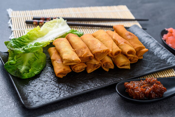 Fried spring rolls with hot sauce and pickled ginger on black slate plate on dark stone slate background.