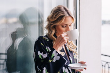 Attractive girl with long hair in pajama having breakfast on balcony in the morning in city. She holds a cup and smiling to camera