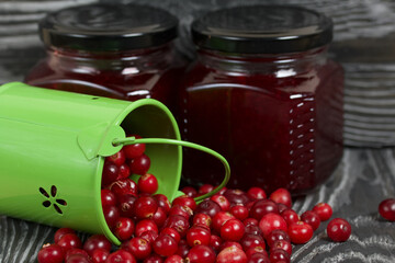 Red cranberries in an overturned green bucket. Lies on brushed boards. Next to it is cranberry jam in a jar.