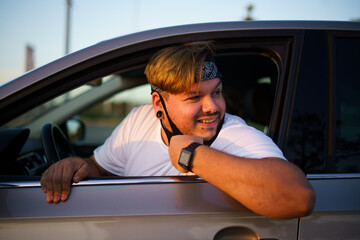 Naklejka premium Chico joven conduciendo su coche recien lavado y comprobando los espejos y cinturones usando mascarilla de la pandemia de coronavirus covid-19