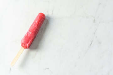 A single bright red popsicle with wooden handle rest on a white and gray marble countertop with...