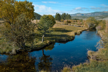 Fototapeta na wymiar Bright blue winding river flows in grass, bushes, trees among the mountains and blue sky with clouds. Reflections in the water. Autumn landscape