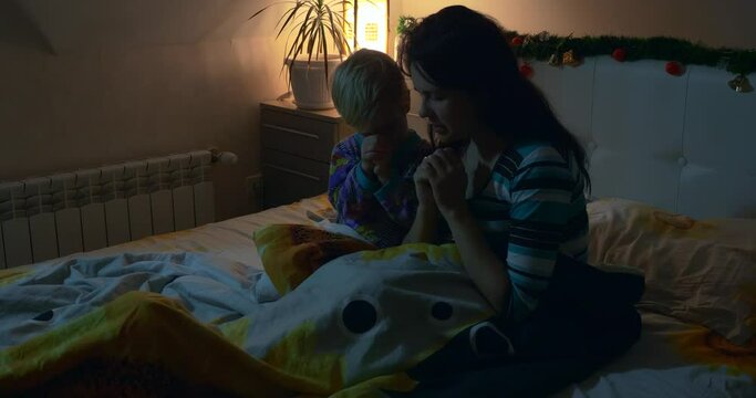 Night Prayer in Bedroom. Mother and Child Praying Sitting on Bed Before Sleeping. Christmas Eve Evening. New Year Holidays