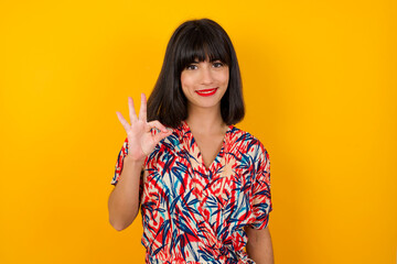 Glad attractive woman shows ok sign with hand as expresses approval, has cheerful expression, being optimistic. Standing against white wall.