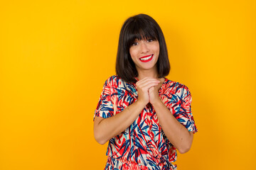 Dreamy charming young female with pleasant expression, keeps hands crossed near face, excited about something pleasant, poses against gray background.