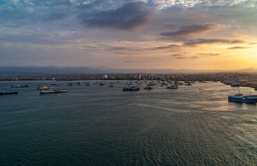 Fototapeta premium Manta, Ecuador - December 2, 2008: Sunset over harbor shows dark ocean water up front with many small boats on top, light over cityscape as central belt. Cloudscape and mountain range in back.