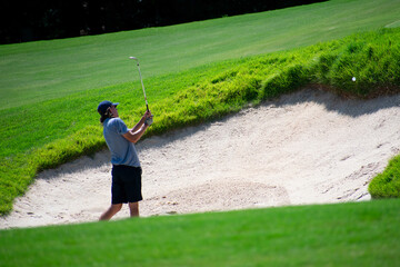 A man golfer chipping a golf ball out of a sand bunker to a green with a pin using a sand wedge at a golf course country club