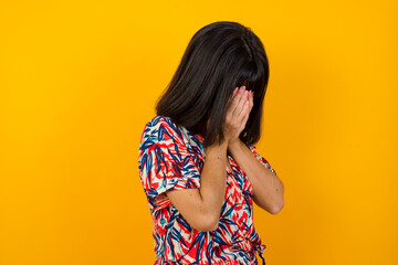 Sad young Caucasian woman wearing casual clothes standing against yellow background covering his face with hands and crying.