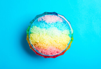 Rainbow shaving ice in glass dessert bowl on light blue background, top view