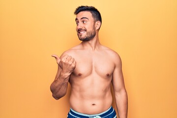 Fototapeta premium Young handsome man with beard wearing sleeveless t-shirt standing over yellow background smiling with happy face looking and pointing to the side with thumb up.