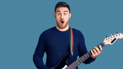 Young handsome man playing electric guitar scared and amazed with open mouth for surprise, disbelief face