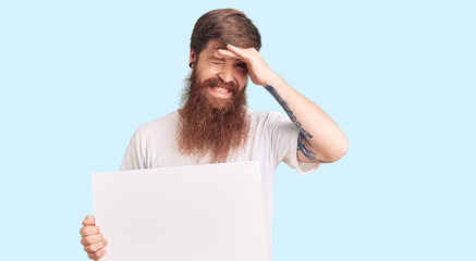 Handsome young red head man with long beard holding blank empty banner stressed and frustrated with hand on head, surprised and angry face