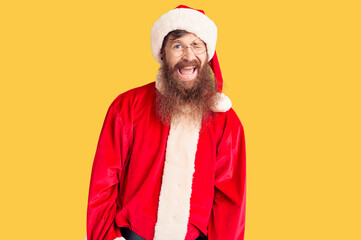 Handsome young red head man with long beard wearing santa claus costume winking looking at the...