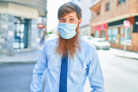 Young handsome redhead businessman wearing coronavirus protection medica mask standing at street of city.
