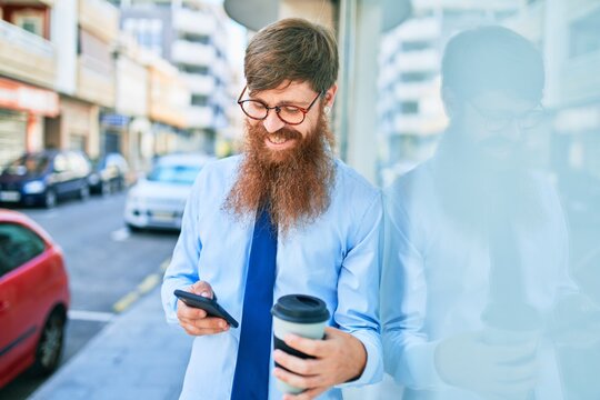 Young handsome redhead businessman smiling happy. Leaning on the wall with smile on face using smartphone drinking take away coffee at street of city.