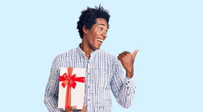 Handsome african american man with afro hair holding gift pointing thumb up to the side smiling happy with open mouth