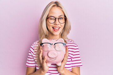 Beautiful caucasian blonde girl holding piggy bank with glasses winking looking at the camera with sexy expression, cheerful and happy face.