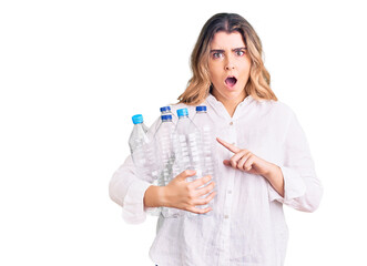 Young caucasian woman holding recycling plastic bottles scared and amazed with open mouth for surprise, disbelief face - obrazy, fototapety, plakaty