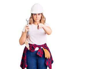 Young caucasian woman wearing hardhat holding wrench pointing with finger to the camera and to you, confident gesture looking serious