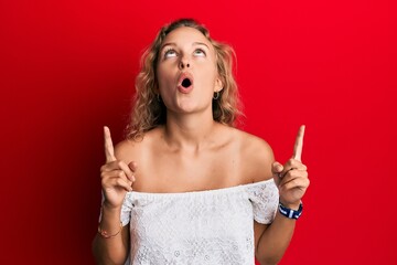 Beautiful caucasian woman wearing casual clothes over red background amazed and surprised looking up and pointing with fingers and raised arms.