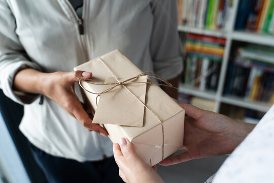 Close up on hands of unknown caucasian woman holding wrapped gift box birthday present handing to the other female person at home or office in day - giving and receiving concept