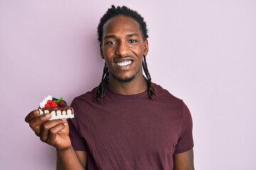 African american man with braids holding chocolate cake looking positive and happy standing and smiling with a confident smile showing teeth