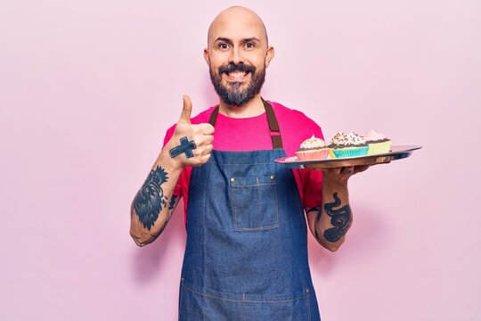 Young handsome man wearing apron holding cupcake smiling happy and positive, thumb up doing excellent and approval sign