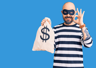 Young handsome man wearing burglar mask holding money bag with dollar symbol doing ok sign with fingers, smiling friendly gesturing excellent symbol