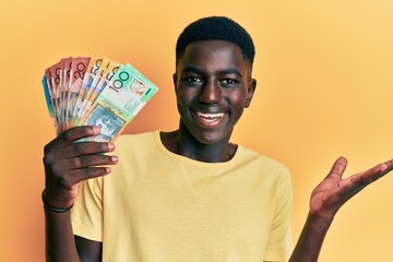 Young african american man holding australian dollars celebrating achievement with happy smile and winner expression with raised hand