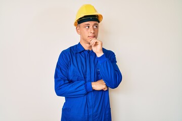 Young hispanic boy wearing worker uniform and hardhat serious face thinking about question with hand on chin, thoughtful about confusing idea