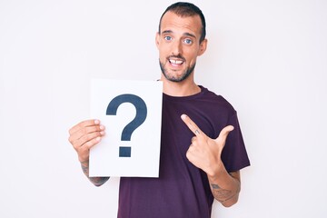 Young handsome man with tattoo holding question mark smiling happy pointing with hand and finger