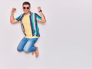 Young handsome hispanic man wearing casual clothes and sunglasses smiling happy. Jumping with smile on face celebrating with fists up over isolated white background